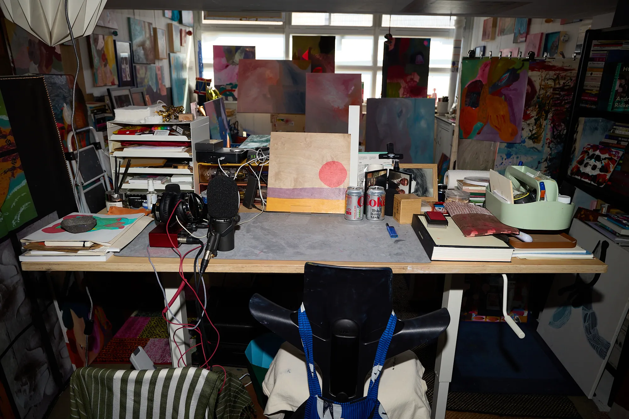 A cluttered desk in Omar Kholeif's studio, with a microphone, headphones, notebooks and paintings propped against the window