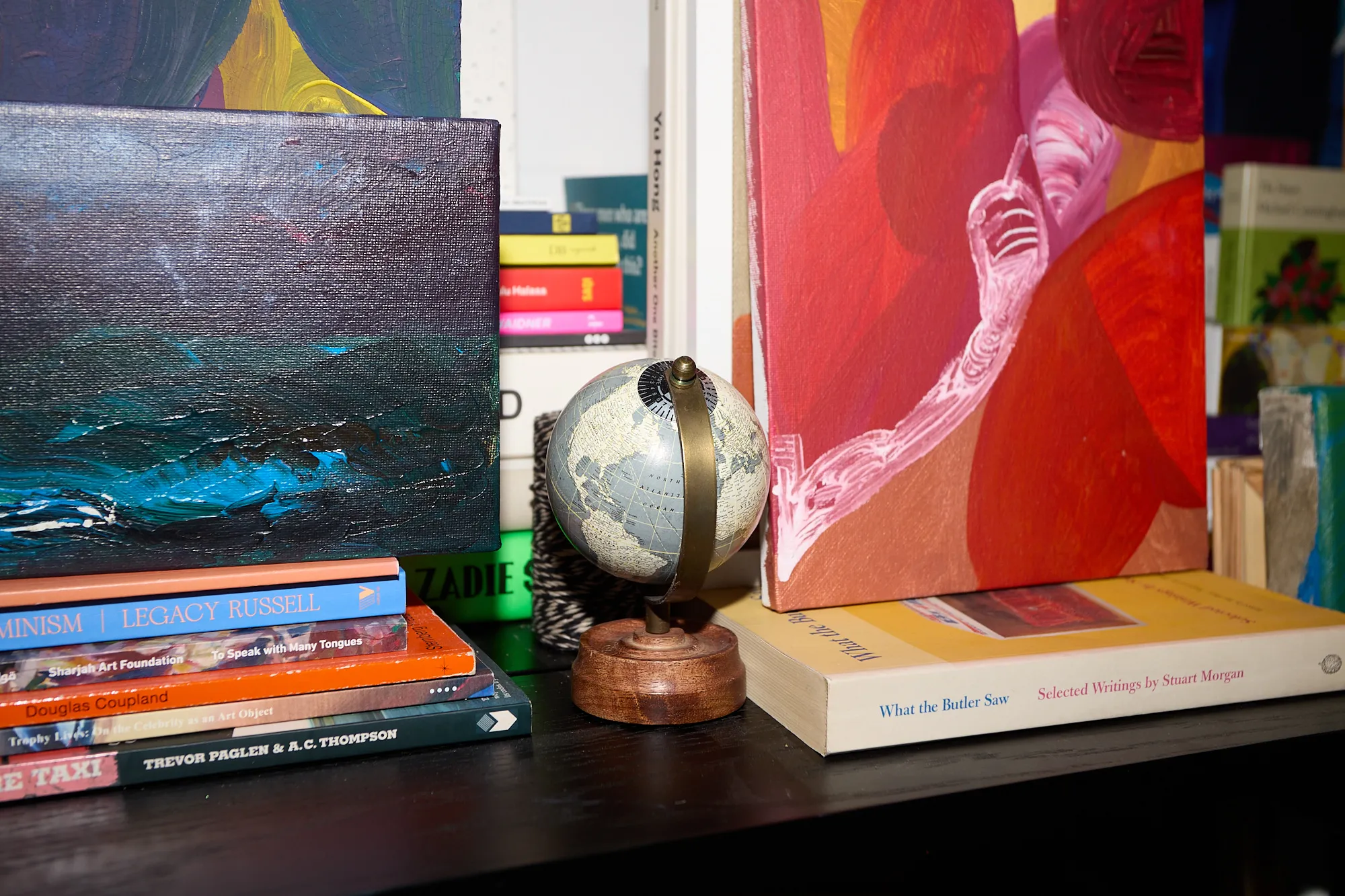 A small globe and stacked art books beside framed paintings on a shelf in Omar Kholeif's studio