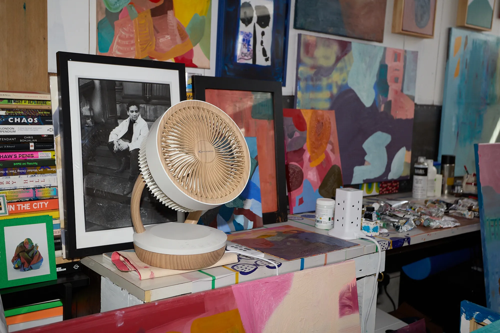 A desk fan and a framed photograph in front of a wall of paintings in Omar Kholeif's studio
