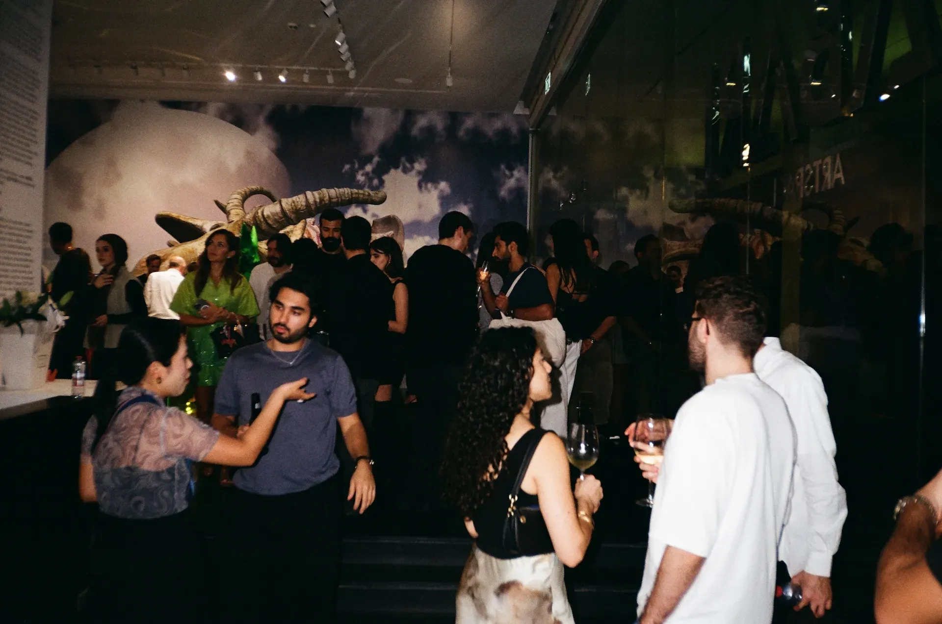 A crowded gallery room with guests talking beneath a large sculptural skull