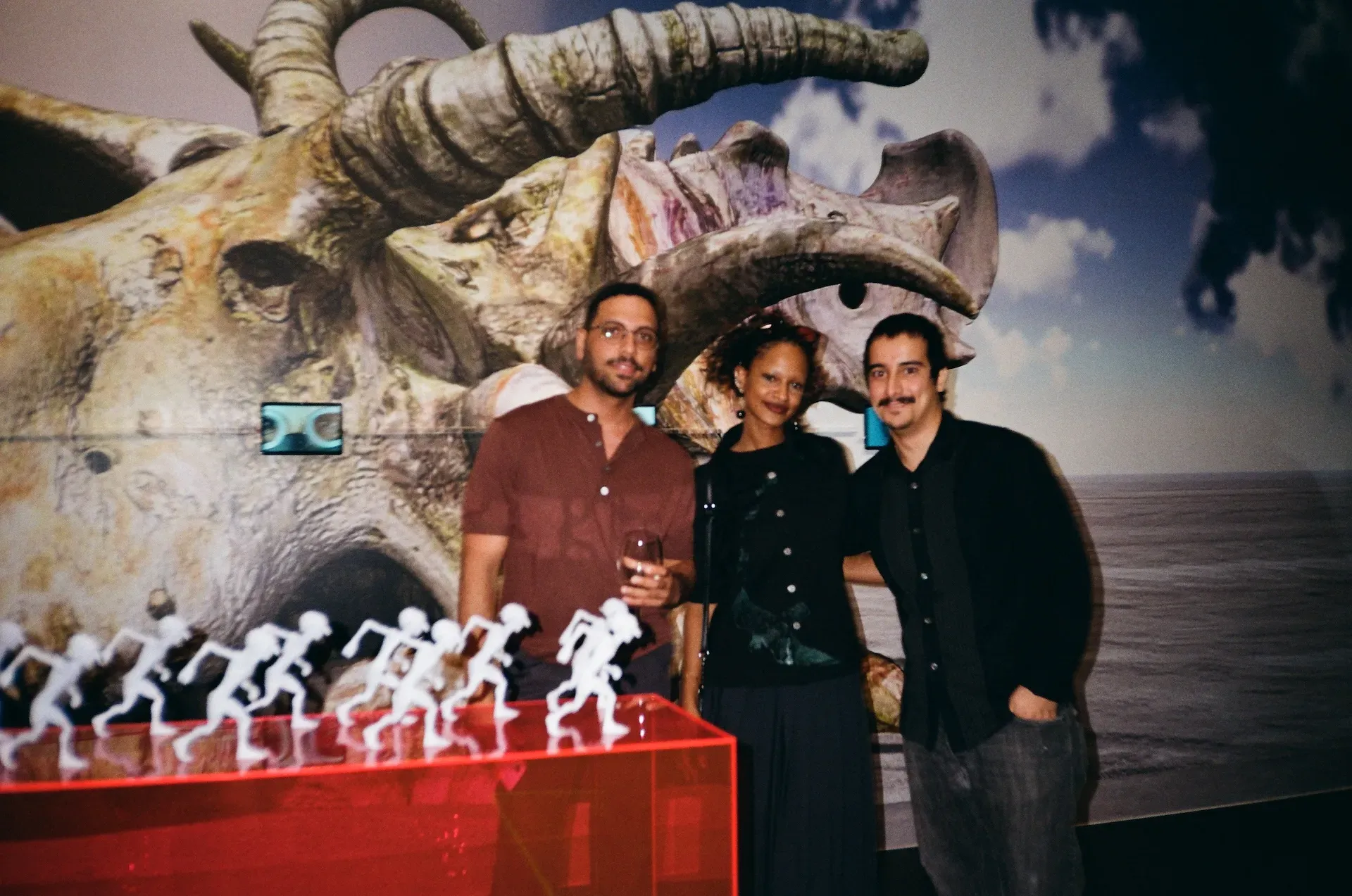 Three guests posing in front of a large sculptural skull and a row of small running figurines in a red display case